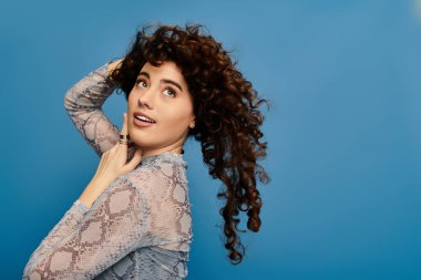 Emotional woman with curly hair poses playfully by a blue wall, capturing a stylish moment. clipart