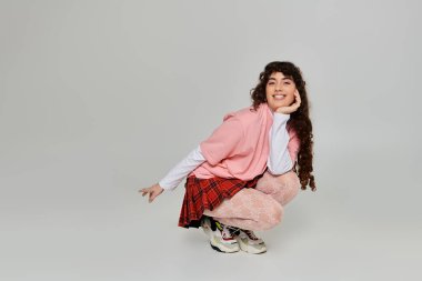 Kıvırcık saçlı genç bir kadın, moda anlayışını fotoğraf çekiminde neşeli bir pozda sergiliyor..