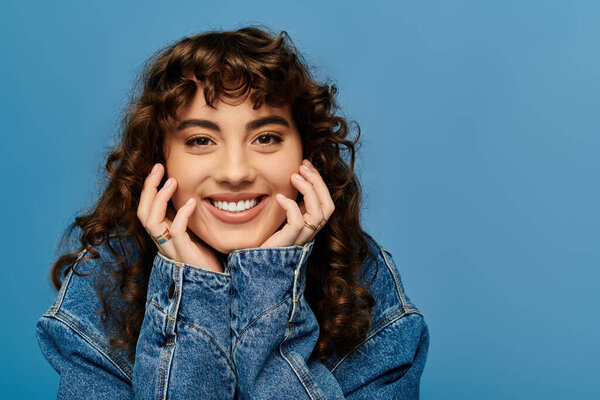Стильная женщина радостно позирует с яркой улыбкой на синем фоне.