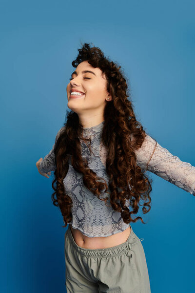 A young woman with long curly hair joyfully spreads her arms, showcasing her trendy outfit.