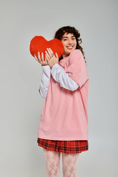 Молодая женщина с вьющимися волосами сияет от радости, держа в руках красную подушку сердца, одетую в модный наряд.