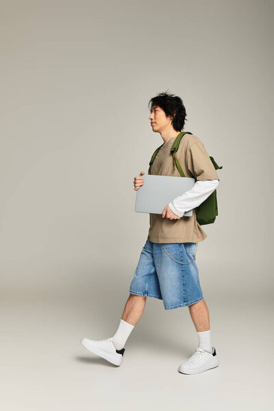 A handsome young asian man is strolling in a modern urban environment while carrying a laptop.