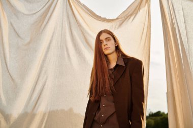 Uzun saçlı genç adam günbatımında dalgalanan kumaşla şık bir kıyafet modelliyor..