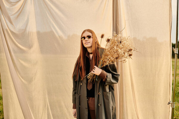 A stylish young man with long hair holds dried flowers against a backdrop of flowing fabric.