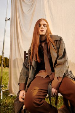 Uzun saçlı genç bir adam kendine güvenerek poz veriyor, sakin bir açık hava ortamında şık giyiniyor..