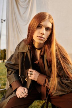 Uzun saçlı bir genç gün batımında çimenlerde diz çökerken şık giyinir..