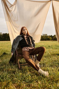 Uzun saçlı genç bir adam gün batımında kumaş perde önünde zahmetsizce poz verir..