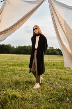 Uzun saçlı şık giyinmiş bir adam huzurlu bir gökyüzünün altındaki yemyeşil bir tarlada geziniyor..