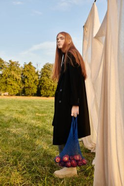 Şık bir genç adam bir çanta elma tutuyor, düşünceli bir şekilde sakin bir manzaraya bakıyor..