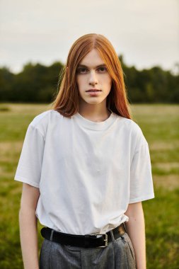 Uzun saçlı genç bir adam, doğanın ortasında kendine güvenen şık bir kıyafetle poz verir..