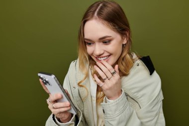 Şık giyinmiş genç bir kadın telefonuna bakarken mutlu bir şekilde gülümsüyor..