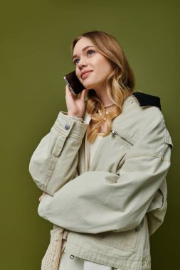 Moda giyinmiş genç bir kadın, yeşil bir fon karşısında telefonla konuşurken gülümsüyor..