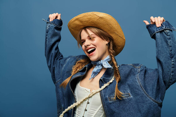 Радостная молодая женщина в джинсовой куртке и соломенной шляпе выражает свое волнение в студии.