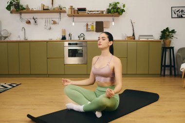 Genç atletik bir kadın paspasın üzerinde oturmuş şık mutfağında meditasyon yapıyor..