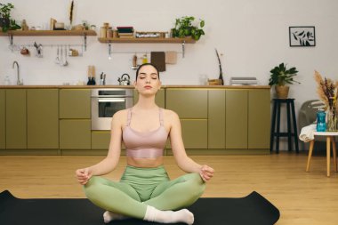 Sportif genç bayan bitkilerle dolu şık bir mutfakta yoga minderinde huzur içinde meditasyon yapıyor..