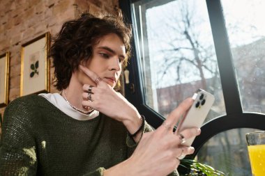 Genç bir adam akıllı telefonuyla parlak gün ışığında büyüleyici bir kafede rahatça iletişim kuruyor..