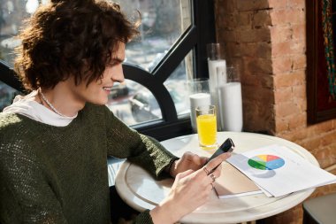 Kafe masasında oturan yakışıklı genç adam akıllı telefonunu yakınlarda bir yerde içerken gülümsüyor..