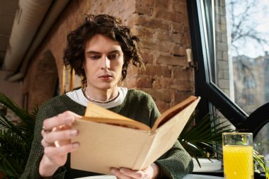 Genç yakışıklı bir adam kafeterya masasında oturuyor, kitap okumaya dalmış, yanında da içki var..