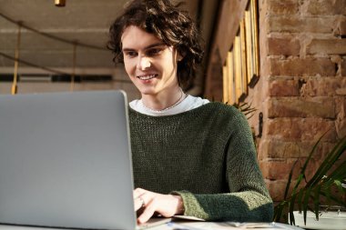 Genç yakışıklı bir adam şık bir kafede oturuyor, çalışırken gülümsüyor ve laptopuna odaklanıyor..