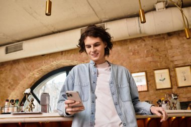 Genç yakışıklı bir adam bir kafenin tezgahına yaslanmış, telefonunun ve içkisinin keyfini çıkarıyor..
