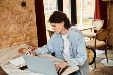 Genç bir adam ilginç bir kafede dizüstü bilgisayarına odaklanırken dikkatlice bir deftere yazar..