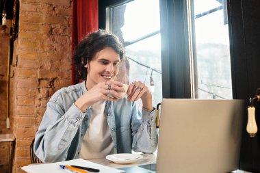 Genç yakışıklı bir adam rahat bir kafede kahveyi yudumlarken gülümsüyor, dizüstü bilgisayarıyla meşgul..