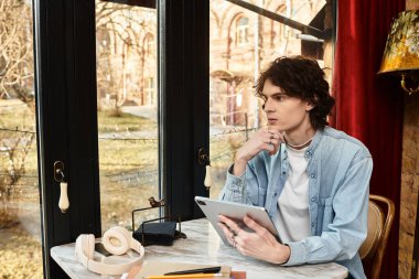 Genç bir adam kafeterya masasında düşünceli bir şekilde oturuyor, tabletine dalmış, dışarıda huzurlu bir manzarası var..