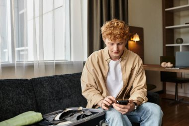 Genç bir adam otel odasında telefonuna bakarak rahatlıyor..
