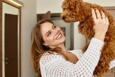 Güzel genç bir kadın, sıcak bir otel ortamında tüylü köpeğiyle oynarken neşeyle gülümsüyor..