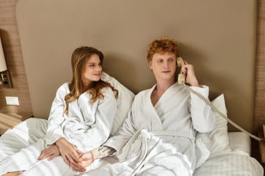 Otel cüppeli genç bir çift telefonda konuşurken yatakta birlikte gülümsüyor..
