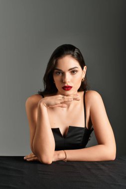 Genç bir kadın, kırmızı dudaklarıyla kendine güveni ve büyüleyici bakışlarıyla gri bir arka plana bakar..