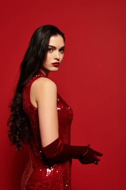 Kahverengi saçlı ve kırmızı dudaklı büyüleyici genç bir kadın çarpıcı kırmızı bir elbise içinde zarifçe poz veriyor..