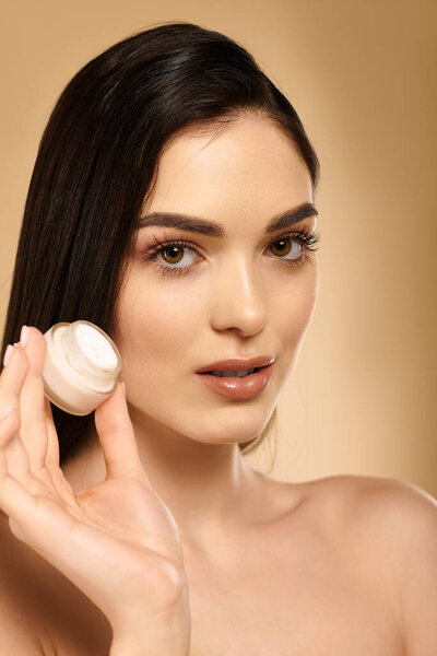 A young woman highlights her glowy, healthy skin while holding a skincare product in a studio.