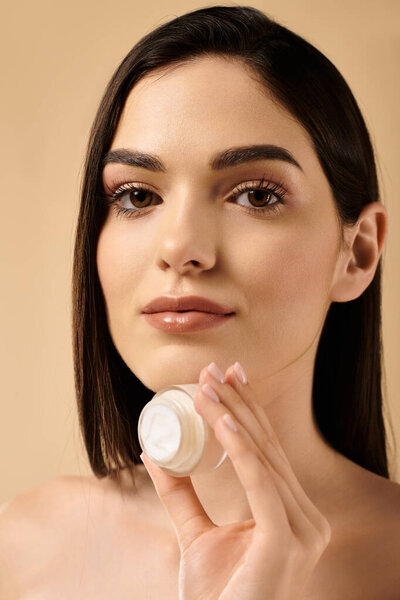 A young woman showcases her flawless skin while holding skincare cream in a studio setting.