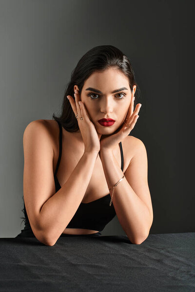 Красивая молодая женщина с каштановыми волосами и красными губами уверенно позирует на сером фоне.