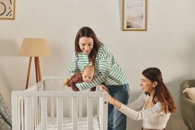 İki anne, sıcak ve davetkar bir oturma odasında bebeklerine neşeyle bakıyorlar..