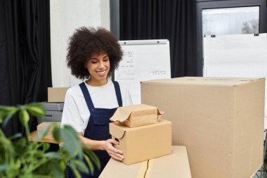 Genç bir kadın, modern bir ofis ortamında kutu ve paketleri düzenlerken gülümsüyor..
