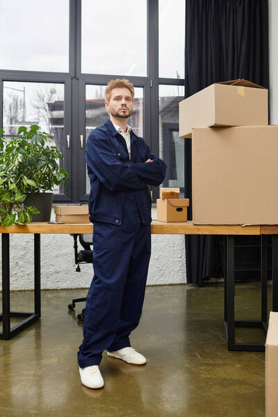 A young man stands confidently in a contemporary office, surrounded by cardboard boxes and plants.
