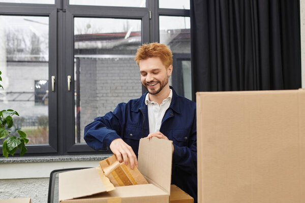 A man happily unpacks a box filled with belongings in a modern room filled with natural light.