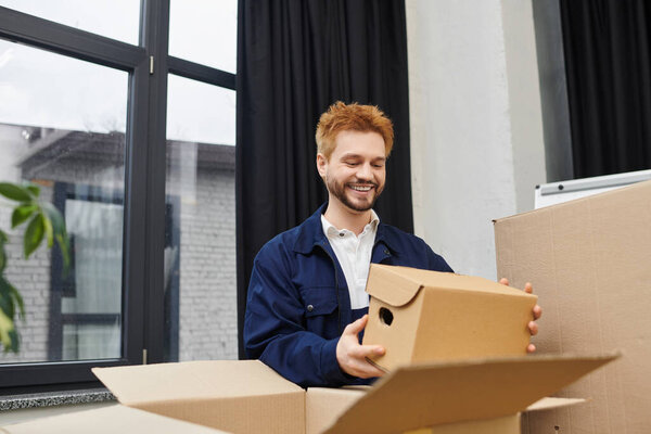 A man smiles happily while unpacking boxes in a modern office setting, surrounded by cardboard.