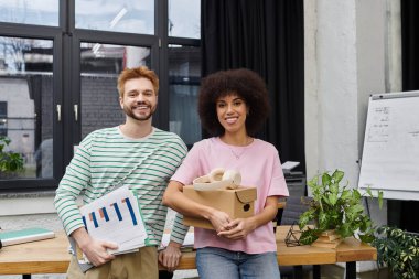 Bitkilerle ve paketlenmiş kutularla çevrili gülümseyen iki birey, taşınma heyecanının tadını çıkarıyorlar..
