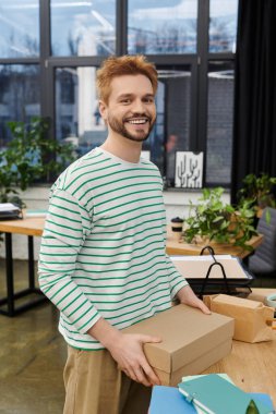 Neşeli bir adam güvenli bir şekilde yeşillikle süslenmiş çağdaş bir ofis alanına kutu koyar..