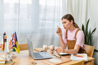 Bayraklar ve notlarla çevrili, parlak, modern bir odada, masasında dikkatlice ders çalışan bir kadın..