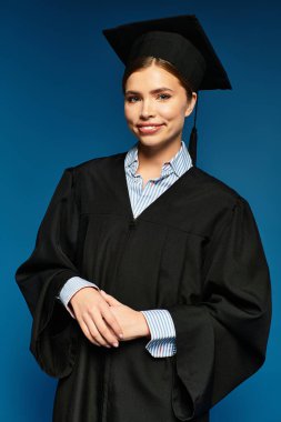 Neşeli genç bir kadın gururla mezuniyet elbisesi ve şapkasıyla canlı mavi bir arka plana poz verir..