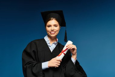 Mezuniyet kıyafetli genç bir kadın, gururla diplomasını mavi arka planda tutuyor..