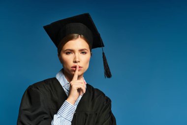Mezuniyet giysisi içinde modern bir genç kadın parmaklarıyla dudaklarını kapatır..