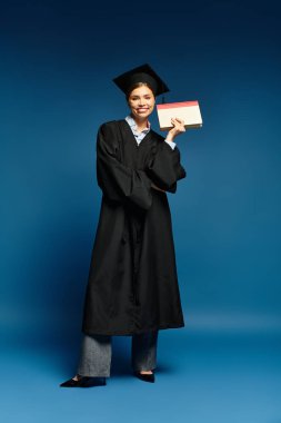 Mezuniyet elbisesi giymiş genç bir kadın mavi bir arkaplanda diplomasıyla gururla gülümsüyor..