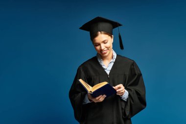 Mezuniyet elbisesi giymiş neşeli genç bir kadın başarılarını yansıtan bir kitap okuyor..