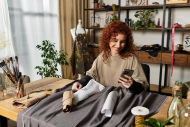 Şık bir çalışma alanındaki kumaş ve aletlerin arasında telefonundaki neşeli zanaatkar kadın..