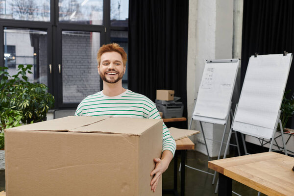 Happy man carries a big box while organizing their workspace for a move.